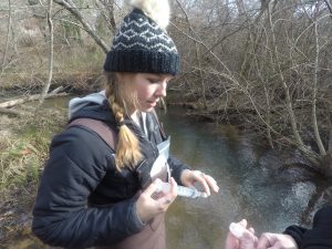 a woman wearing waders and a knit winter hat stands in a river, using a plastic syringe to put a water sample into a small bottle