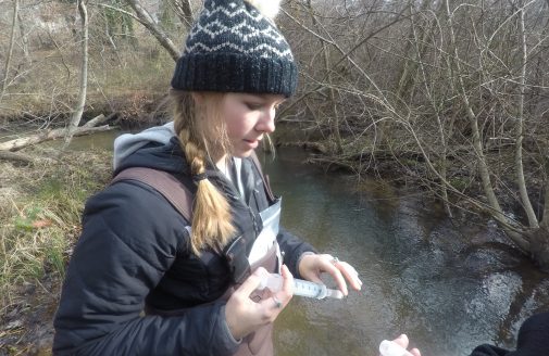 a woman wearing waders and a knit winter hat stands in a river, using a plastic syringe to put a water sample into a small bottle