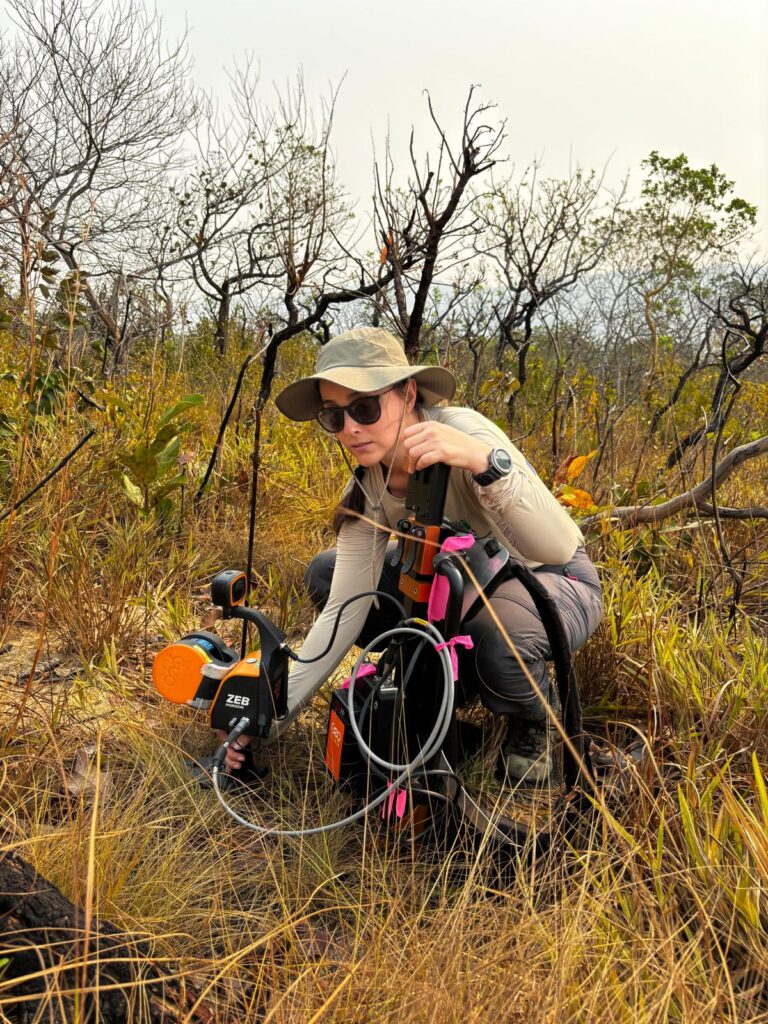 Dr. Machado crouches down in front of some shrubs among brown grass, using a scientific instrument to take a measurement of the ground