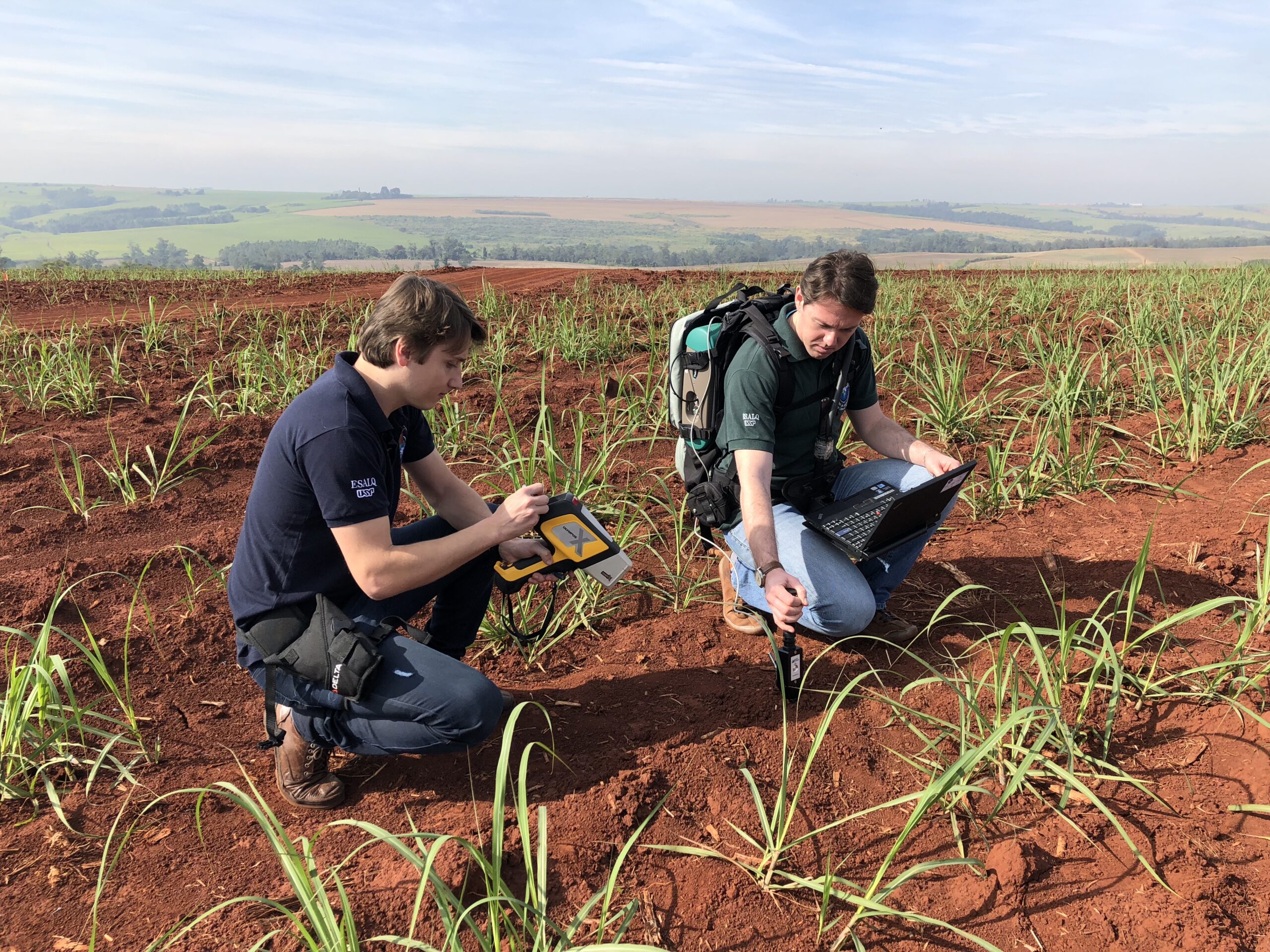 two researchers crouch in an agricultural field, using a handheld scanner to examine the soil