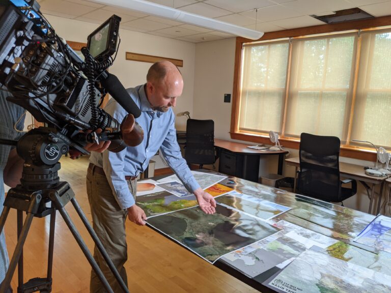 Greg Fiske displays printed out maps on a table to a camera crew