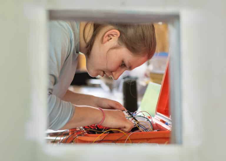 picture through a cut out in an aluminum sheet, Zoe dietrich works on wires in an orange box