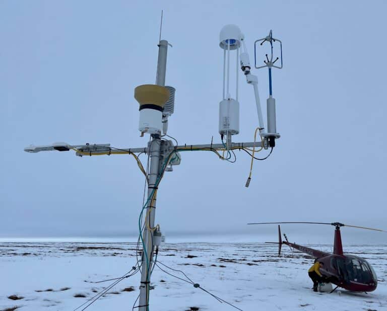 scientific instruments mounted to a slim pole in the foreground, with a helicopter on a snowy field in the background
