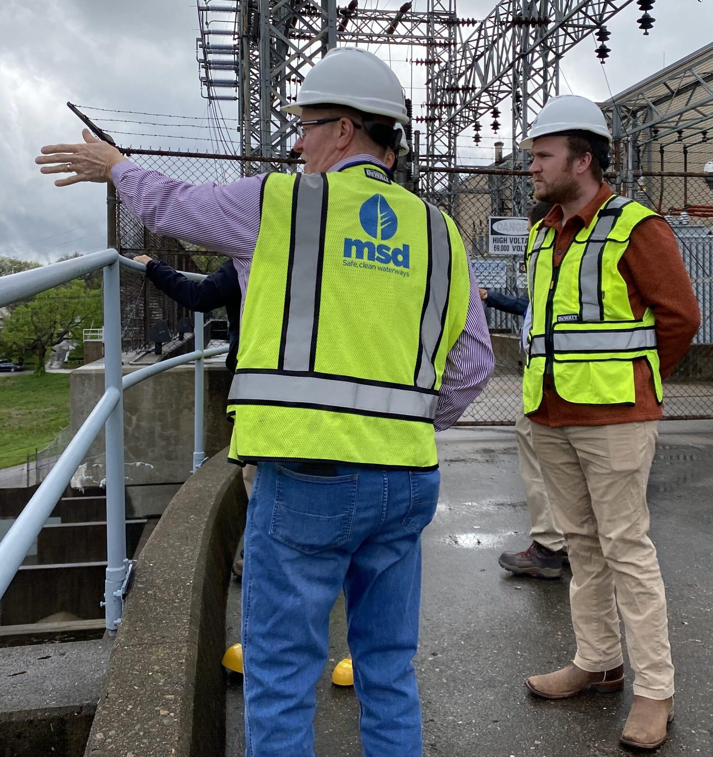two men in hard hats and neon yellow vests look off a balcony with electrical infrastructure in the background