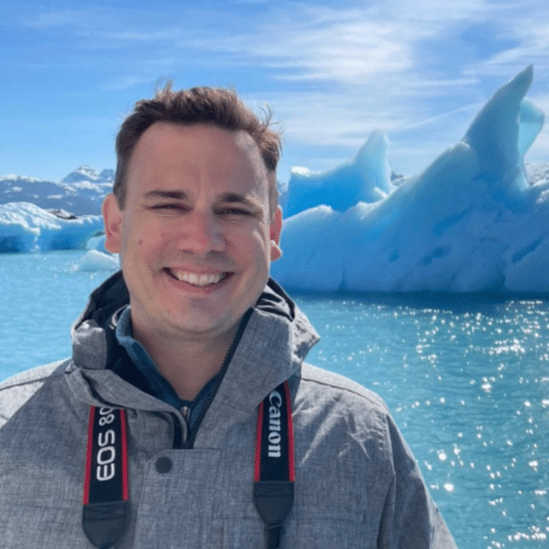 portrait of chris gloninger, a white man with short brown hair, with icebergs and light blue water in the background. he has a camera strap around his neck