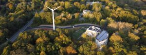 an aerial photograph of the Center building, next to its tall white wind turbine, surrounded by green trees with a golden shine from sunset. The waters of the bay are visible in the background