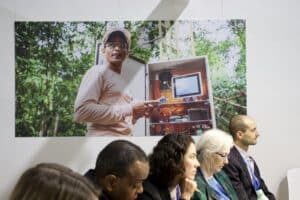 a row of seated people listens to an out-of-frame presentation. On the wall behind them is a large photo of someone demonstrating computer equipment in the forest