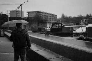 a greyscale photo of a man in a coat standing under an umbrella, facing away from the camera