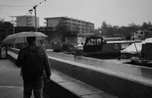 a greyscale photo of a man in a coat standing under an umbrella, facing away from the camera