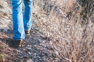 a pair of legs wearing blue jeans and boots walking on a trail next to brown, dormant vegetation