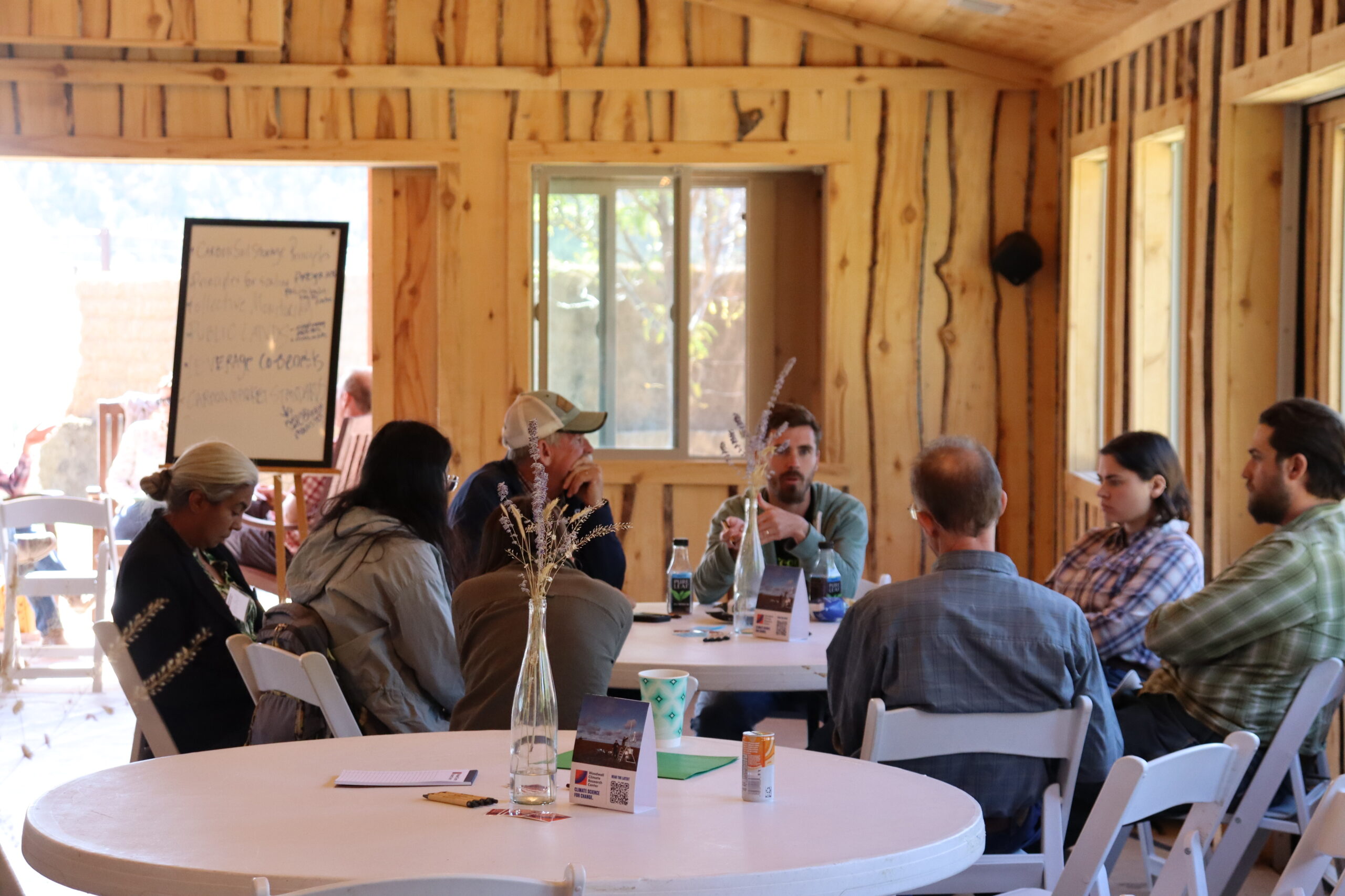 a group of people sits around table having a discussion. the room is well-lit with natural light and wooden walls