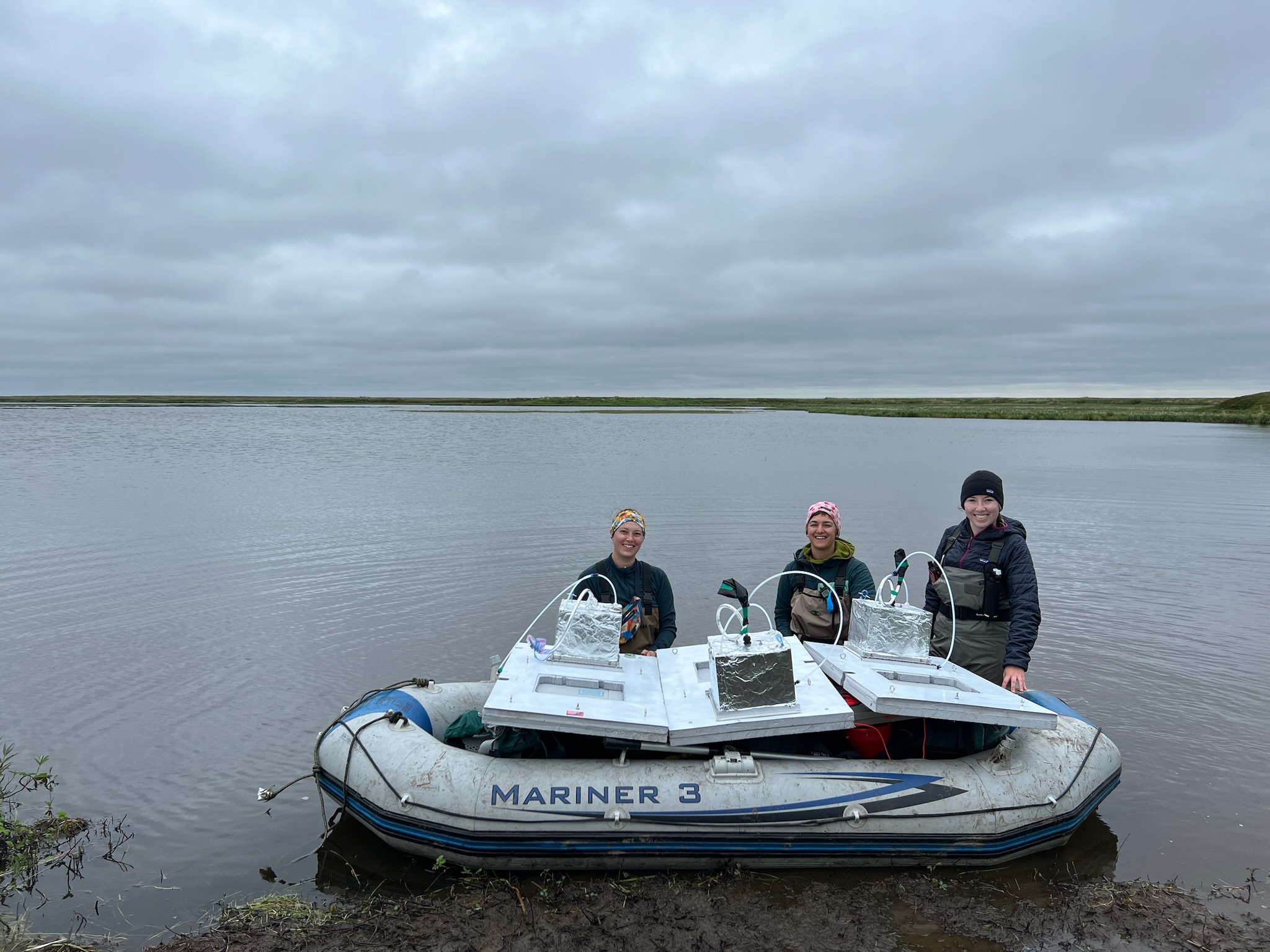 three researchers stand behind a small inflatable boat in the water. on top of the boat are small scientific instruments on flat platforms