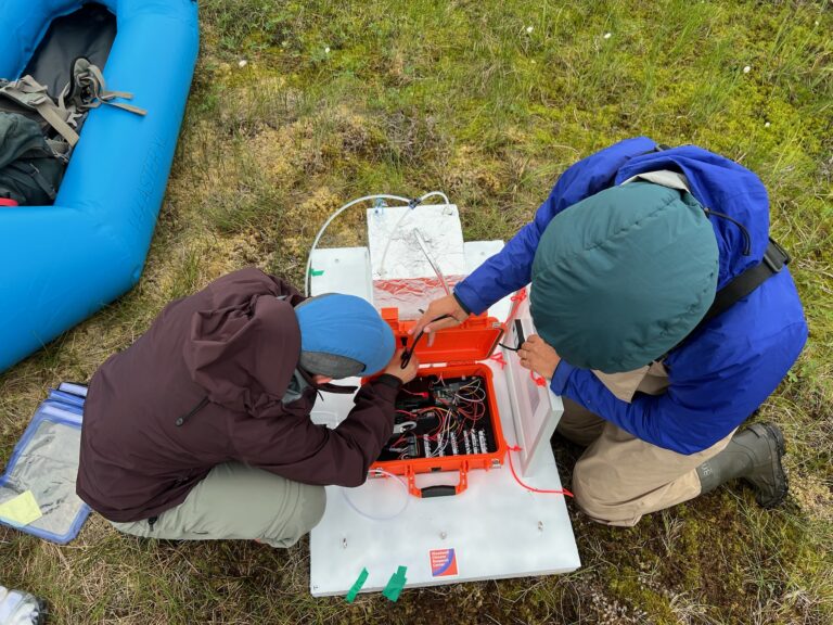 two researchers work on a scientific instrument in an orange plastic case, sitting on the ground on a white flat platform