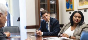 a white man and a Black woman sit at a large wooden table in an office, speaking to someone, with papers lying in front of them