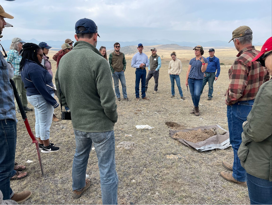 a group of people standing in a circle, looking at a pile of soil on a tarp while one person talks. they are wearing jeans, plaid, and jackets and the grass is very short and dry. mountains are visible in the background