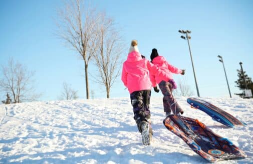 Two children wearing pink winter jackets run up a snowy hill, facing away from the camera, dragging modern toboggan sleds behind them.