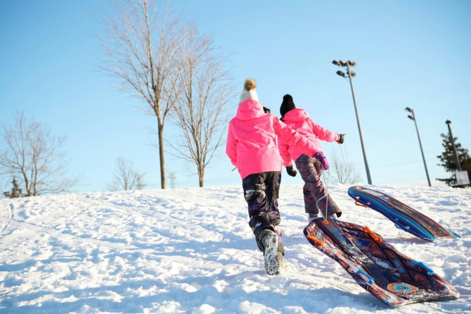 Two children wearing pink winter jackets run up a snowy hill, facing away from the camera, dragging modern toboggan sleds behind them.