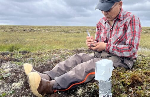 a man sits on the tundra next to a scientific instrument inserted into the ground, taking notes. He is wearing a plaid shirt and a baseball cap