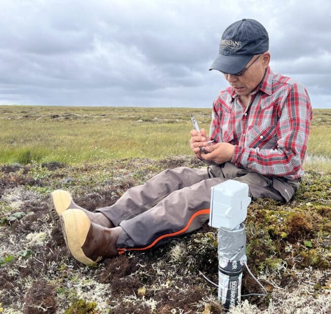 a man sits on the tundra next to a scientific instrument inserted into the ground, taking notes. He is wearing a plaid shirt and a baseball cap