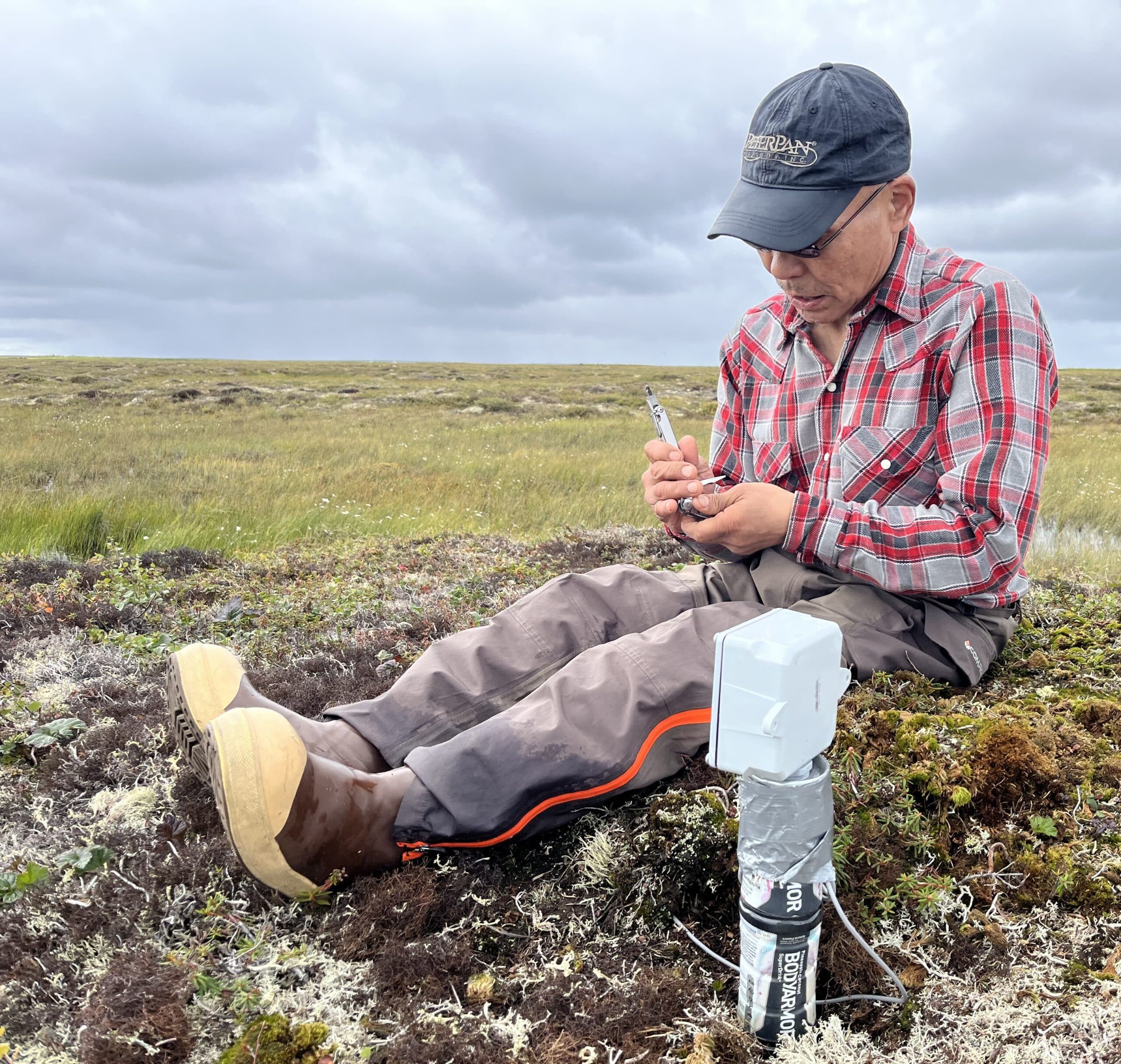 a man sits on the tundra next to a scientific instrument inserted into the ground, taking notes. He is wearing a plaid shirt and a baseball cap