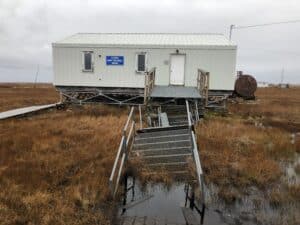 a small building is perched on scaffolding on the tundra, with stairs leading to the building sinking into a puddle of water. the building has a blue sign on it that says "alaska army national guard"
