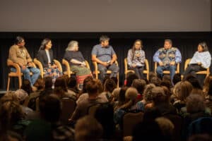 seven people are seated on a stage in front of a seated audience. the panelists include Deb Haaland and Brooke Woods, who is speaking into a handheld microphone