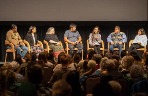 seven people are seated on a stage in front of a seated audience. the panelists include Deb Haaland and Brooke Woods, who is speaking into a handheld microphone