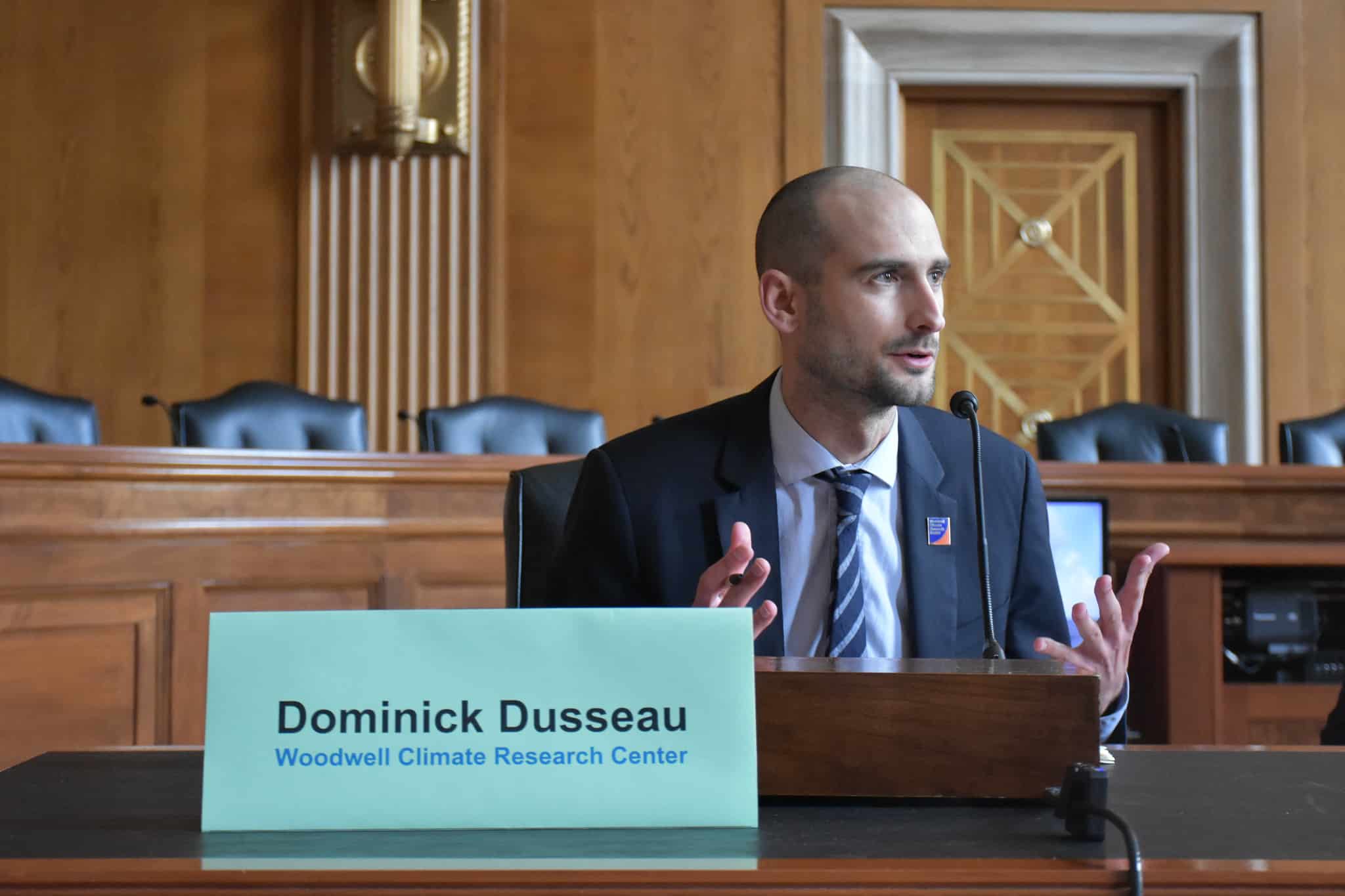 dominick dusseau sits behind a wooden desk in a wood paneled room