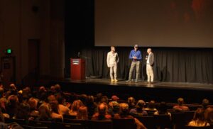 three men holding microphones stand on a stage in front of a projector screen, speaking to a seated audience