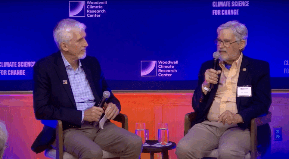 Max Holmes sits next to John Holdren, who is speaking into a microphone