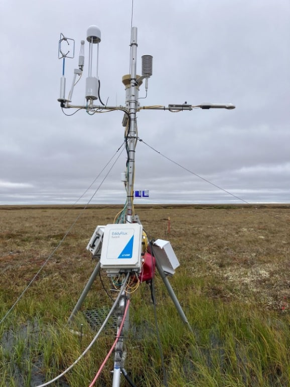 Medium-sized silver pole standing up on three legs in a grassy landscape with a cloudy sky. Small blue containers attached to the middle of the main pole.