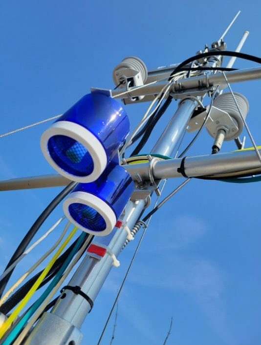 Two blue cylindrical devices attached to a pole with wires hanging down. Blue sky in the background.