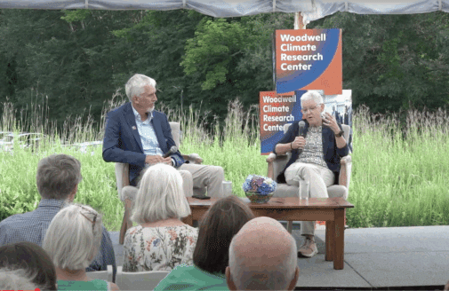 Max Holmes sits next to Gina McCarthy, who is speaking into a microphone to a seated audience
