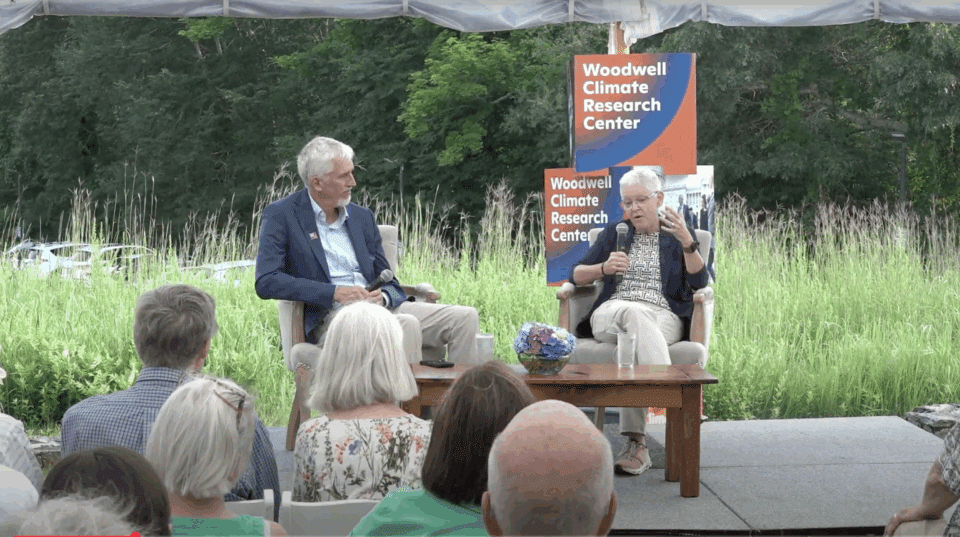 Max Holmes sits next to Gina McCarthy, who is speaking into a microphone to a seated audience