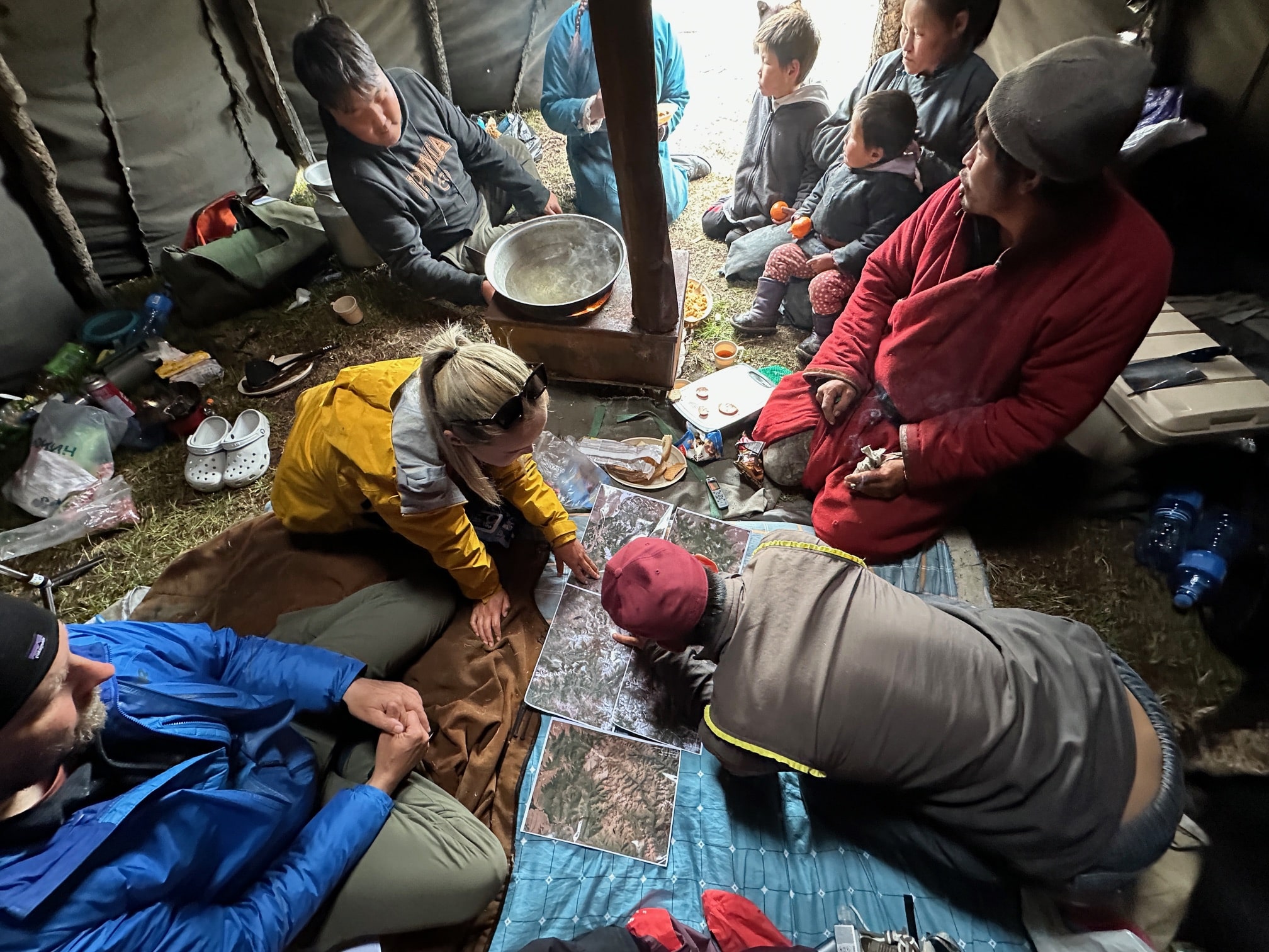 next to a cookstove inside a tent-like structure, people sit on the grass around a printed map, pointing at various features