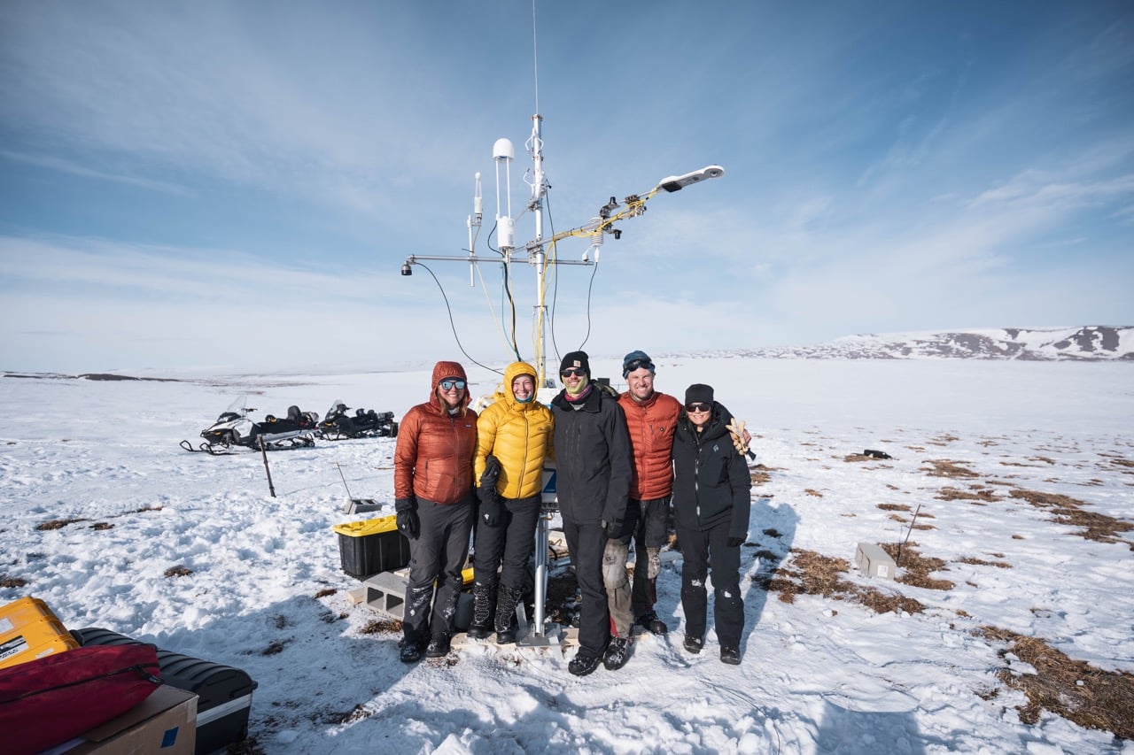5 researchers in bright colored winter coats stand in front of an eddy covariance flux tower on the snowy tundra
