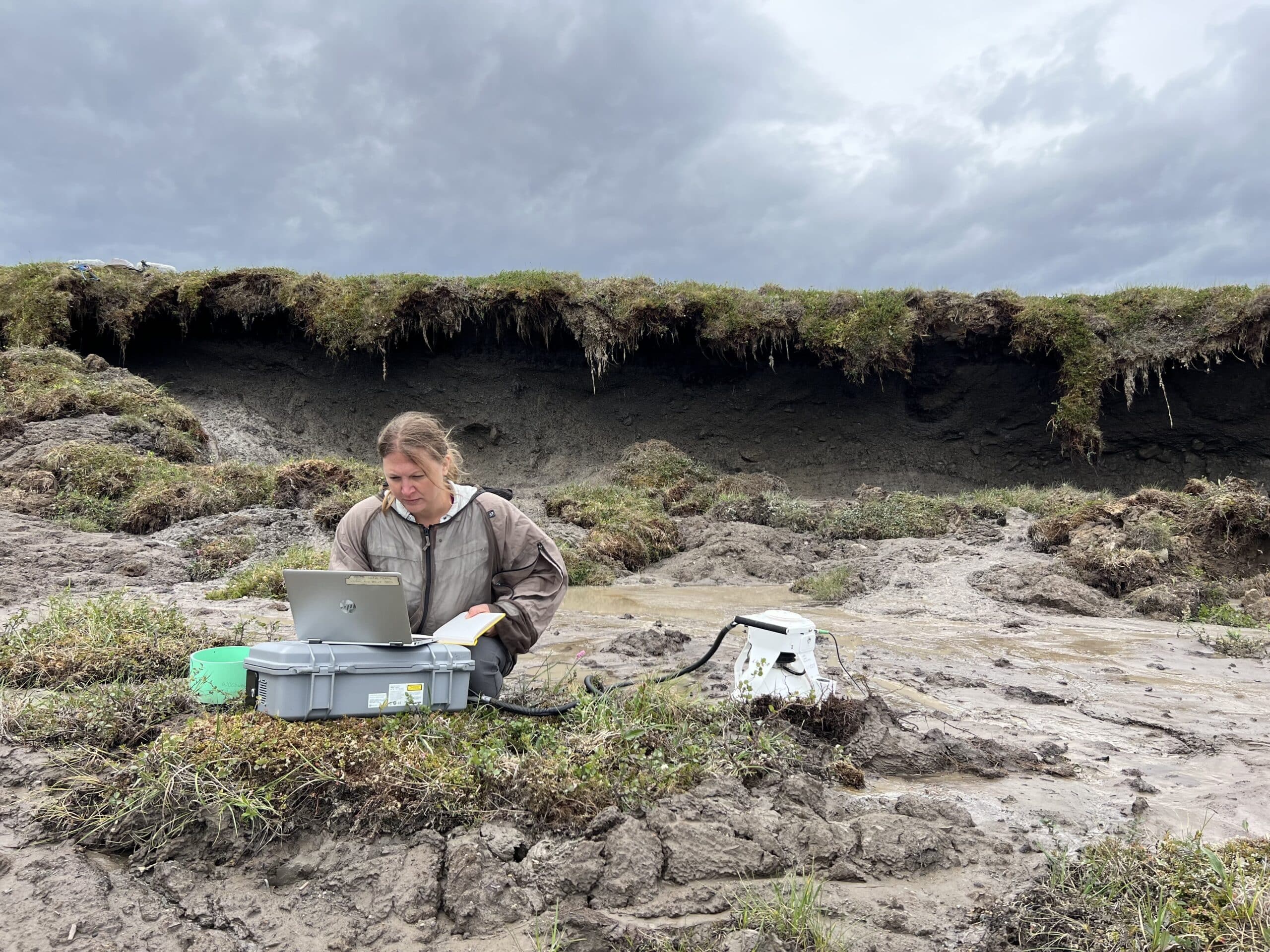 Dr. Jennifer Watts sits on the ground, surrounded by mud and tufts of grass. She is working on a laptop connected to a chamber sitting on the ground next to her. The headwall of the rts is in the background
