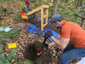 Jonathan Sanderman crouches next to a hole in the forest floor, which he is digging out with a shovel. In the background, loose soil lays on a blue tarp