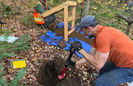 Jonathan Sanderman crouches next to a hole in the forest floor, which he is digging out with a shovel. In the background, loose soil lays on a blue tarp