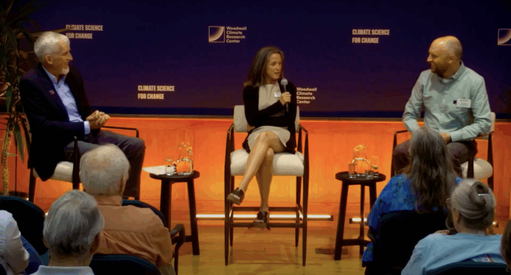 Max Holmes, Sue Natali, and Greg Fiske sit in front of a seated audience while Sue speaks into a microphone