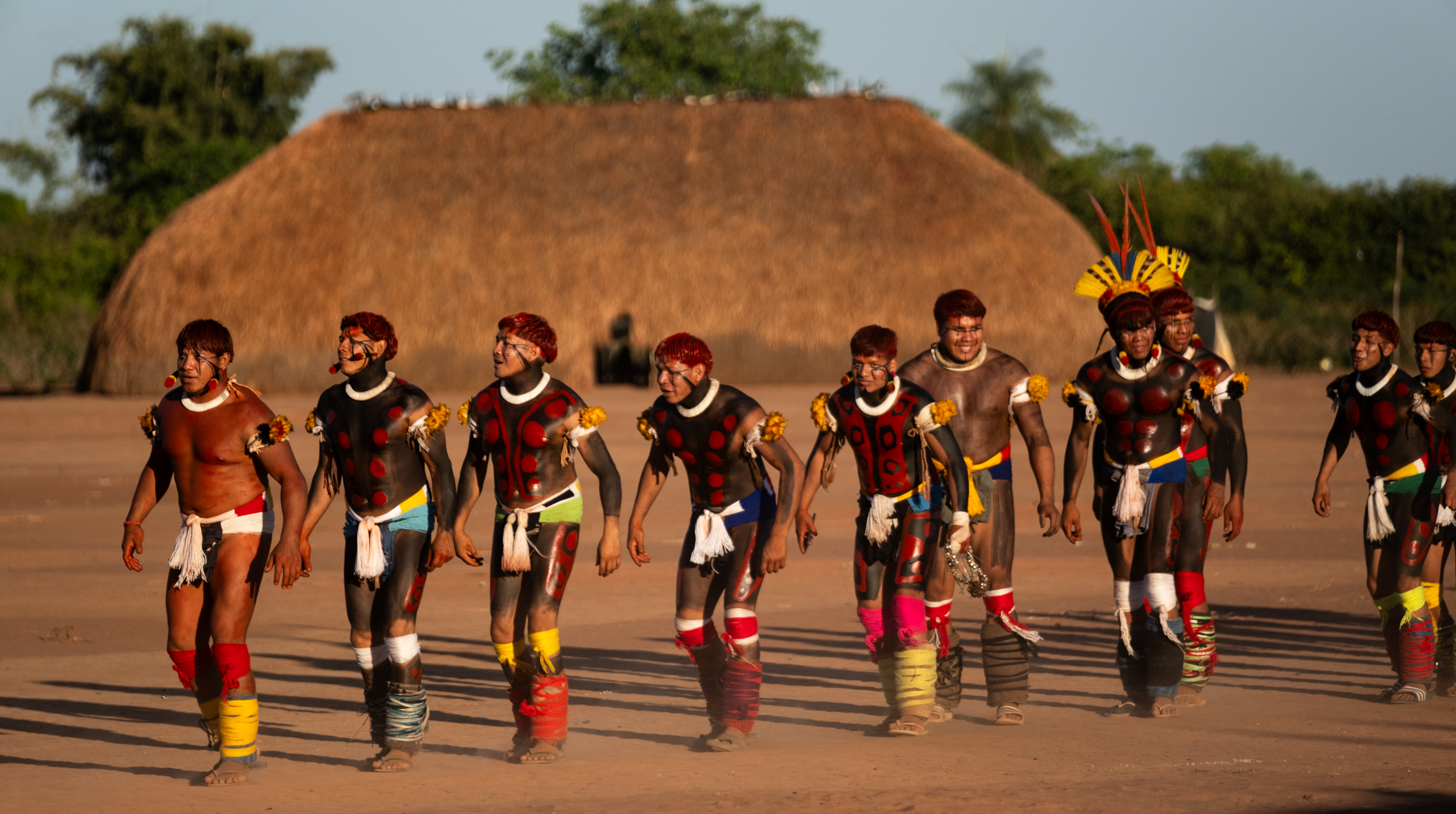 a group of painted dancers in a line in front of a thatched roof stucture