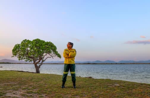 a woman stands with her arms crossed in front of a lake with a single tree in the background