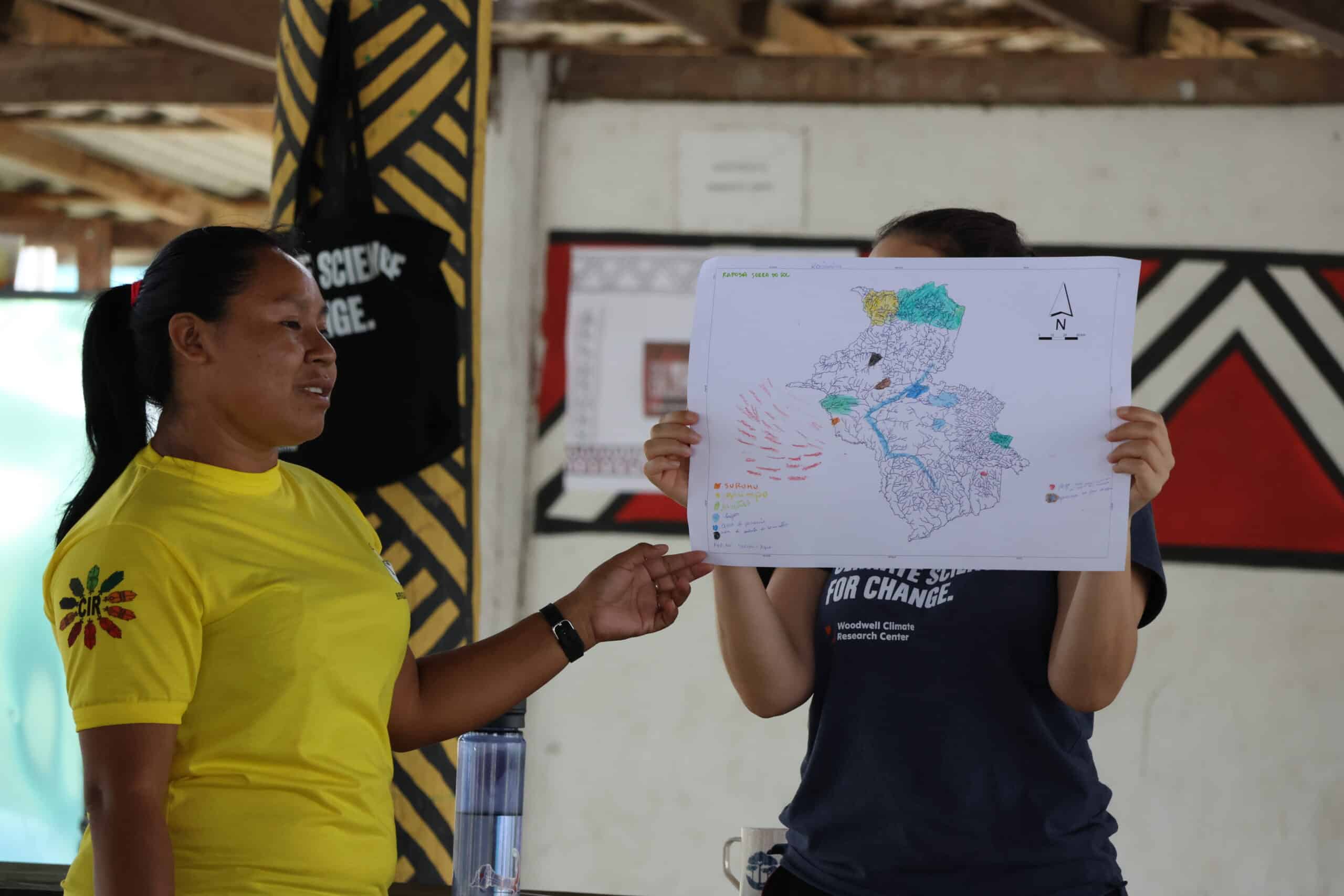 a woman in a yellow shirt points at a hand drawn map held up by manoela machado