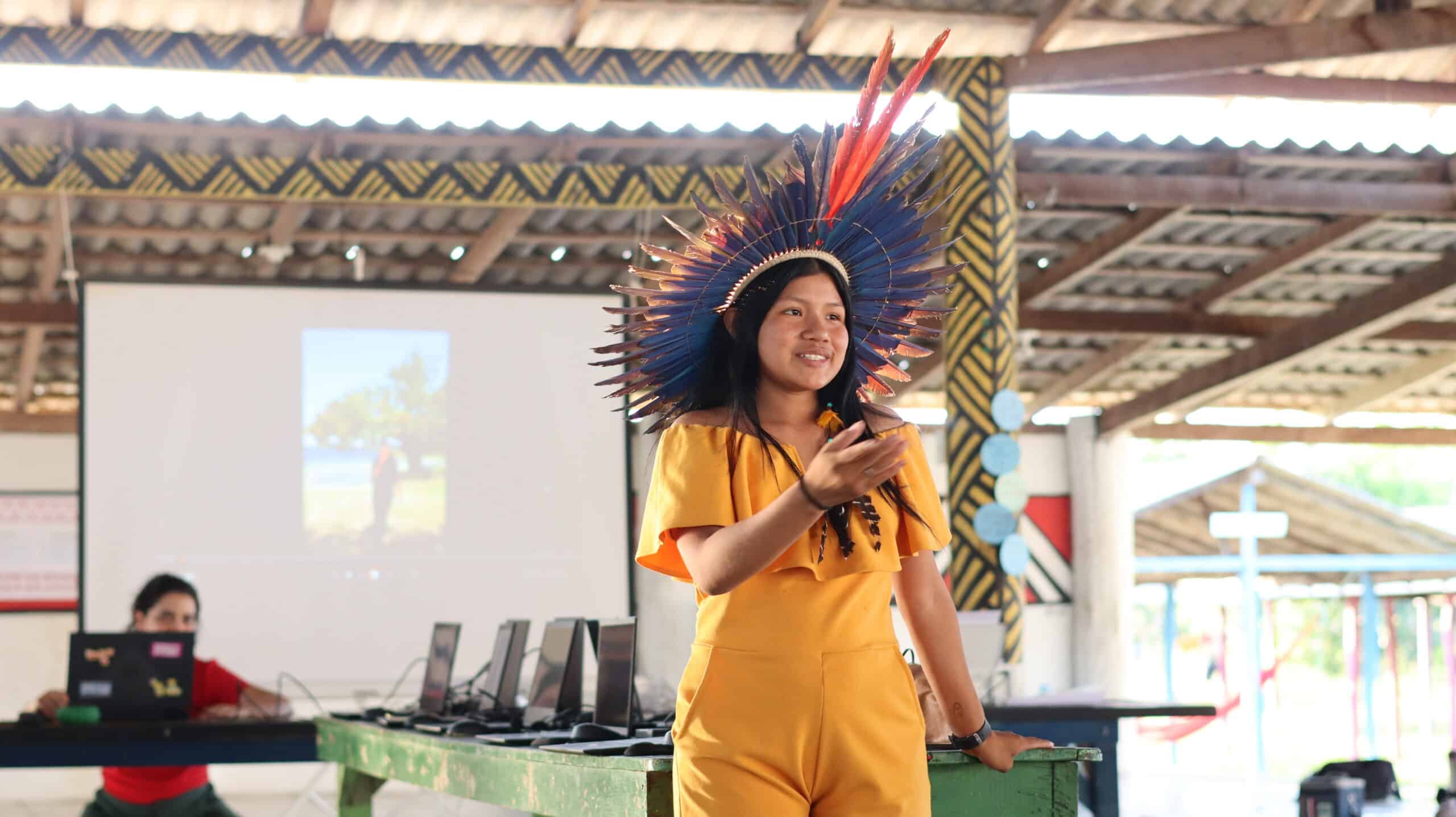 a young woman in a yellow jumpsuit and a headdress of blue and red macaw feathers presents at the workshop in Roraima