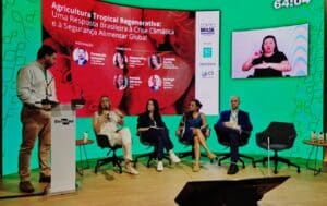 Ludmila Rattis sits on a panel, speaking into a microphone while the others on the panel, and the moderator behind the podium, look at her and listen