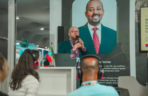 matti goldberg speaks at a podium in front of a poster of the president of ethiopia. people with headsets listen in the foreground