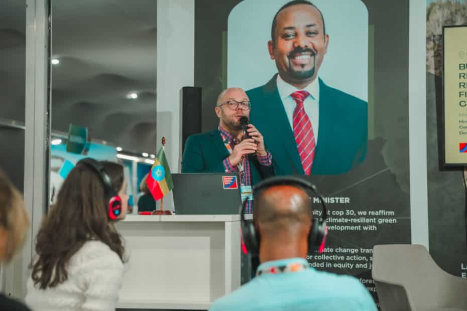 matti goldberg speaks at a podium in front of a poster of the president of ethiopia. people with headsets listen in the foreground