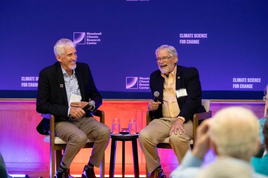 Max Holmes and John holdren sit in front of an audience holding microphones. both men are smiling and laughing
