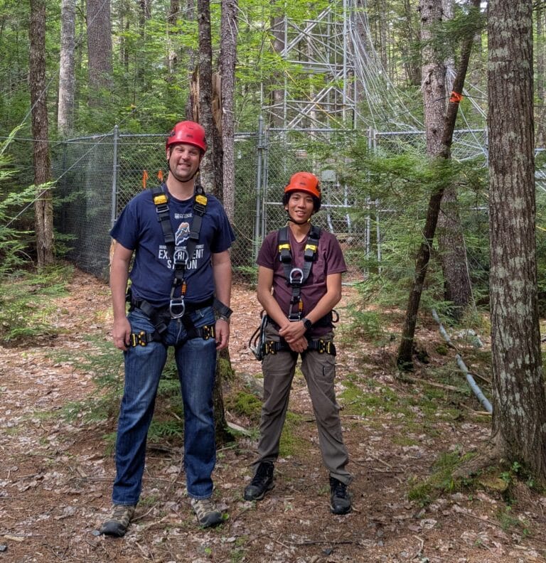 two young people wearing harnesses and orange hard hats stand in the forest, smiling