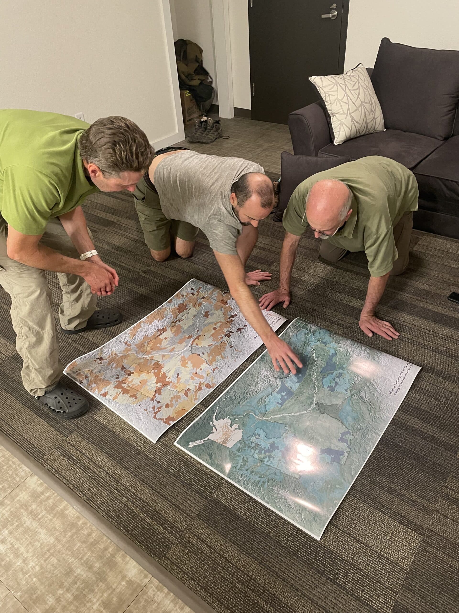 three men kneel on the ground pointing at large print maps of Alaska